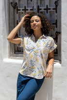 Woman stands in front of a wooden door wearing a neutral based floral cotton top with short folded cuff sleeves, collar and V neckline 