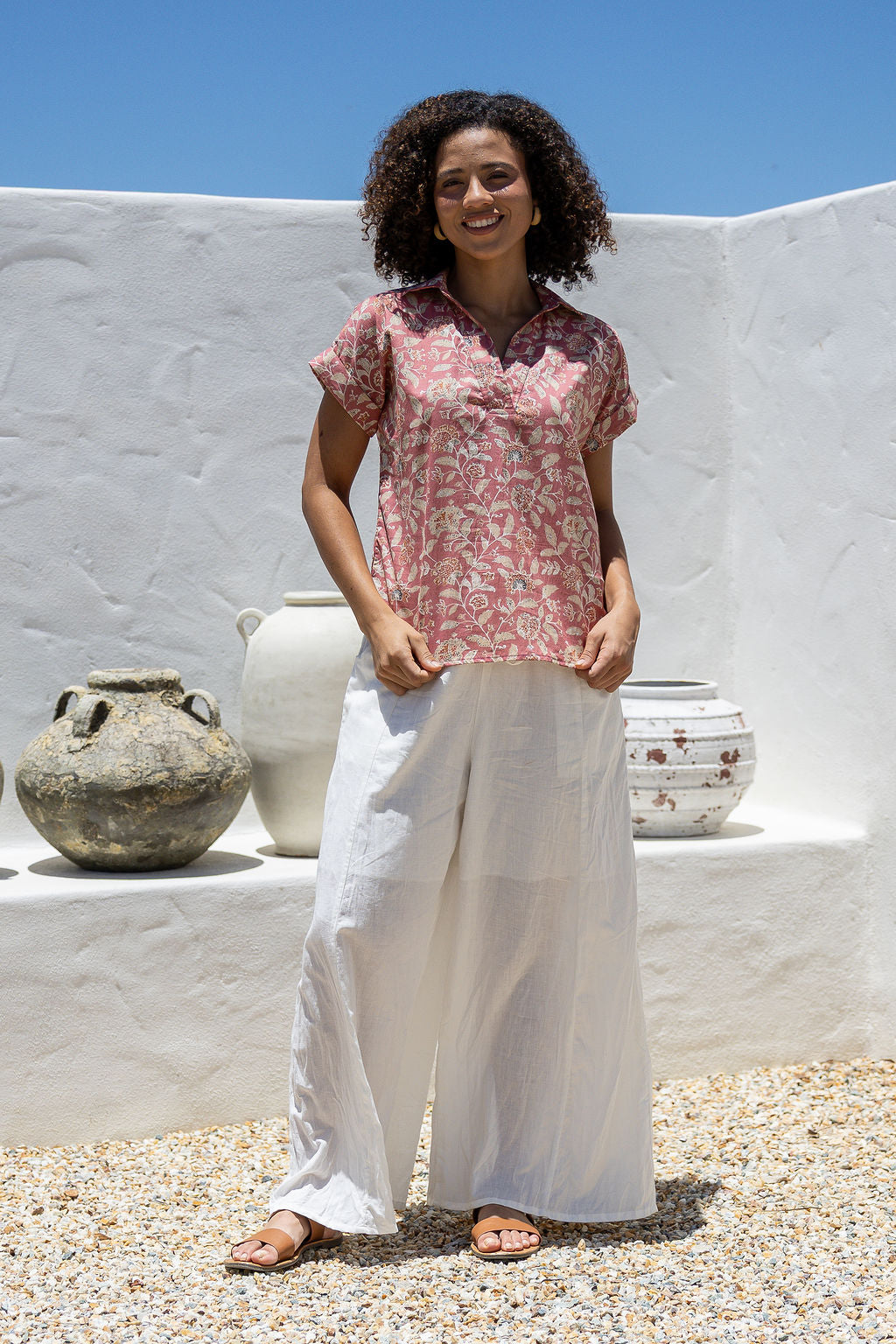 Woman wearing a rose pink based floral 100% cotton top and white pants standing in front of a decorative vases.