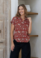Woman stands wearing a red  based floral cotton top with short folded cuff sleeves, collar and V neckline 