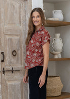 Woman stands sideways wearing a red based floral cotton top with short folded cuff sleeves, collar and V neckline 