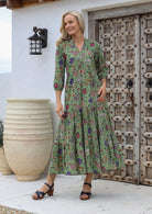 Woman wearing a green floral maxi dress with 3/4 sleeves, buttoned bodice and hands in pocket, standing in front of a rustic wooden door.