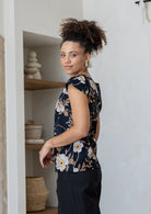 A woman stands sideways wears a 100% cotton tailored top with short cap sleeves and a round neckline with a keyhole cutout, paired with black cotton pants