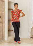 A woman poses in a cap-sleeved cotton tailored top with vibrant red backdrop accented by elegant multicoloured floral vines.