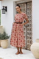 Woman poses with both hand in pockets of short sleeved midlength cotton dress with boat neckline and cream based warm floral print.