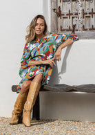 Woman in a colorful dress and tan boots sitting on a bench against a white wall.