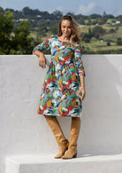Woman in a colorful floral dress and brown boots standing against a white wall with a scenic background.