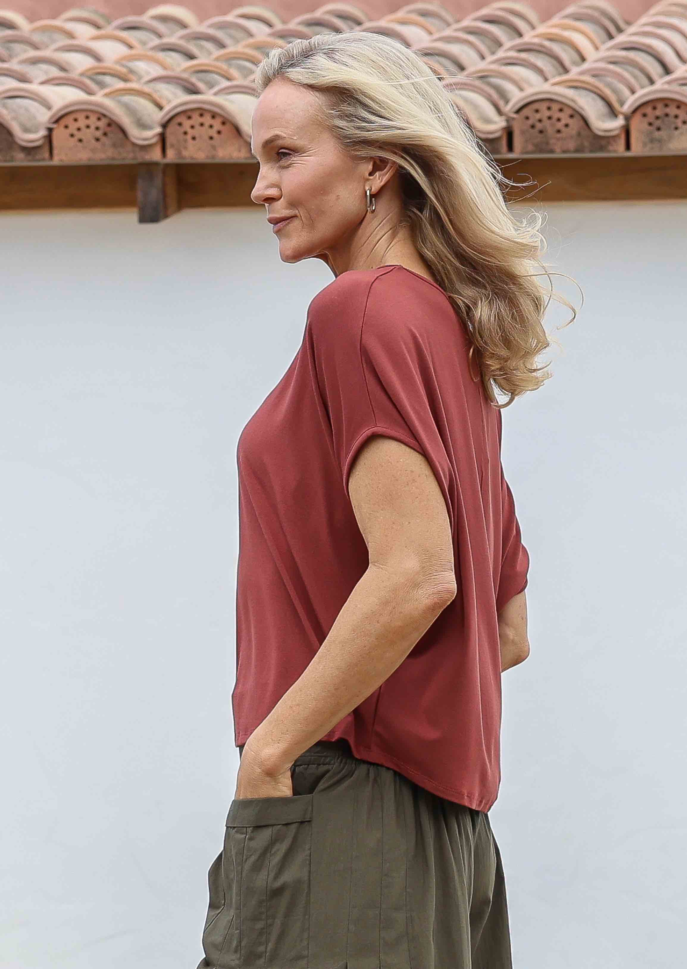 Woman smiling and standing sideways with hand in pocket wearing 100% rayon batwing top in terracotta standing in front of a white house