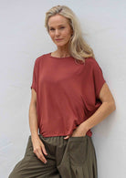 Woman with one hand in pocket wearing 100% rayon batwing top in terracotta standing in front of a white wall 