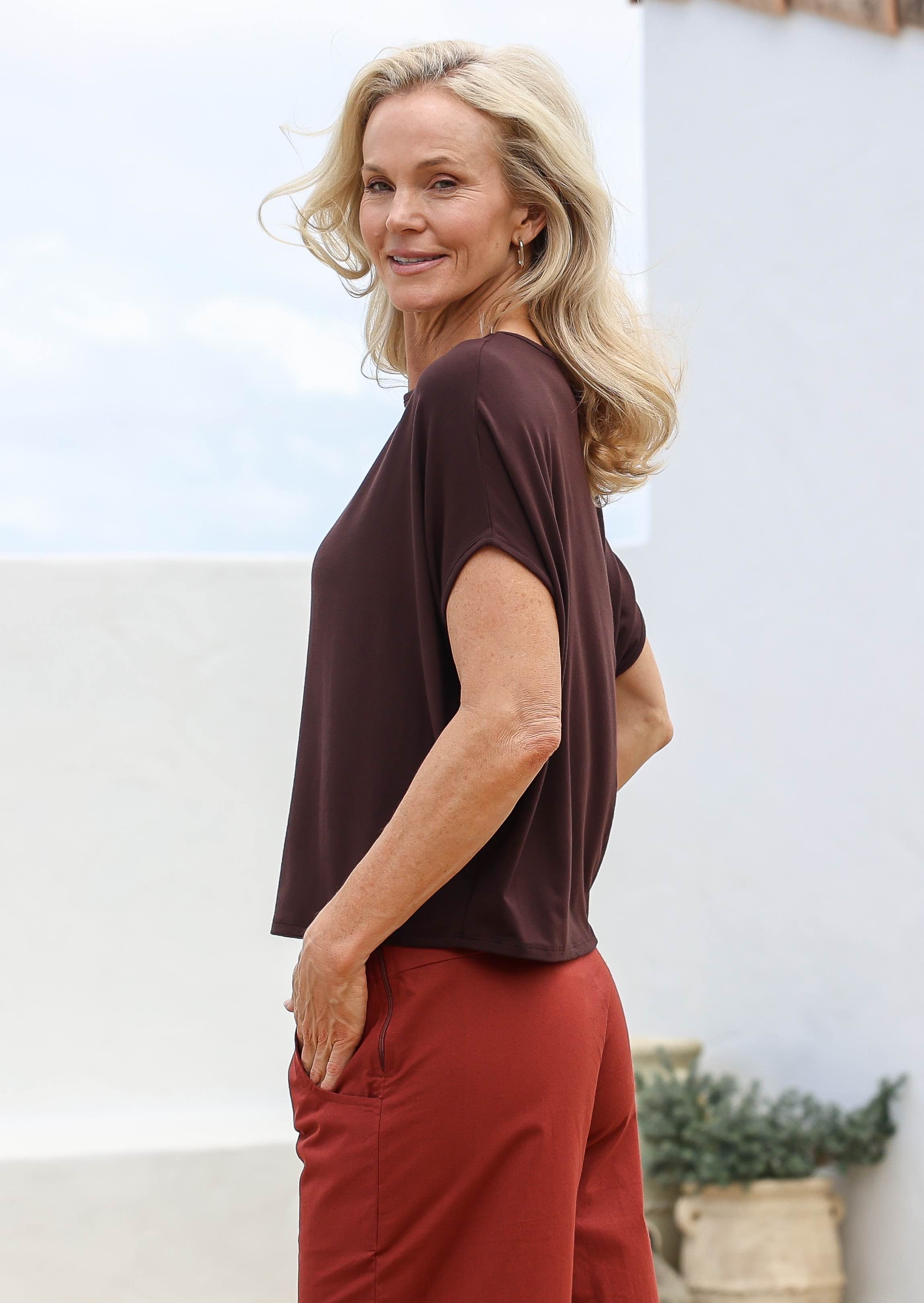 Woman wearing a brown top and burnt henna pants standing outdoors with a light background
