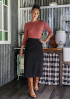 Woman in a dusty pink top and black cotton corduroy midi length A-line skirt standing in a rustic room