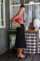 Woman standing side on wearing a black cotton corduroy midi length skirt with hands in back pockets