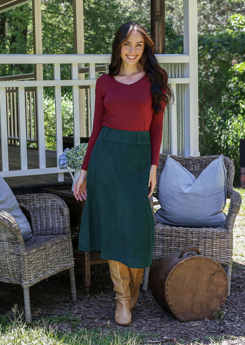 Woman in a red long sleeve top and green cotton corduroy A-line skirt standing near a porch with wicker chairs