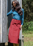 Woman in a teal top and red cotton corduroy midi length skirt standing next to a corrugated metal wall outdoors.