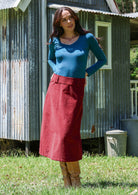 Woman wearing a cabernet red cotton corduroy A-line midi length skirt with belt loops, paired with a teal top and tan boots