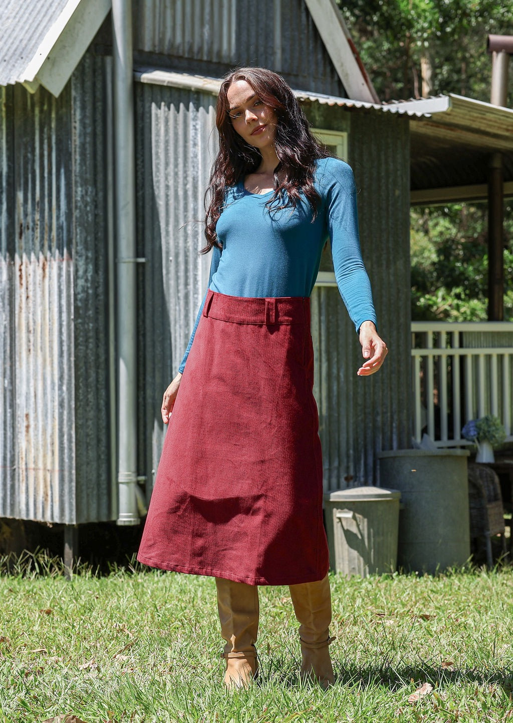 Woman wearing a teal long sleeve top and cabernet red cotton corduroy A-line skirt in front of a rustic building