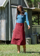Woman wearing a teal long sleeve top and cabernet red cotton corduroy A-line skirt in front of a rustic building