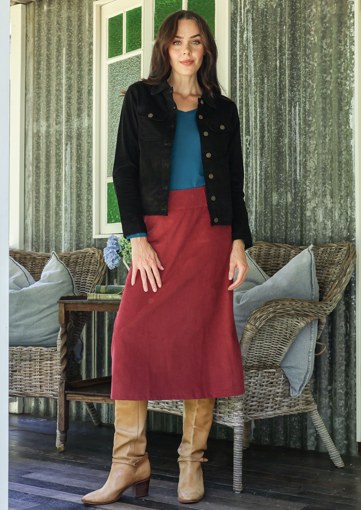 Woman in a red cotton corduroy midi length skirt and black corduroy jacket, paired with a teal top and tan boots