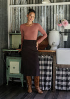 Woman leaning on rustic stove wearing a chocolate brown cotton corduroy A-line midi length skirt paired with a rose pink top