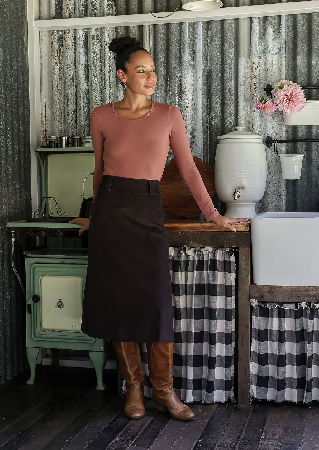 Woman wearing a chocolate brown cotton corduroy A-line midi length skirt paired with a rose pink top and tan boots