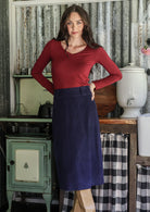 Woman in a maroon top and navy midi length cotton corduroy skirt standing in a rustic room