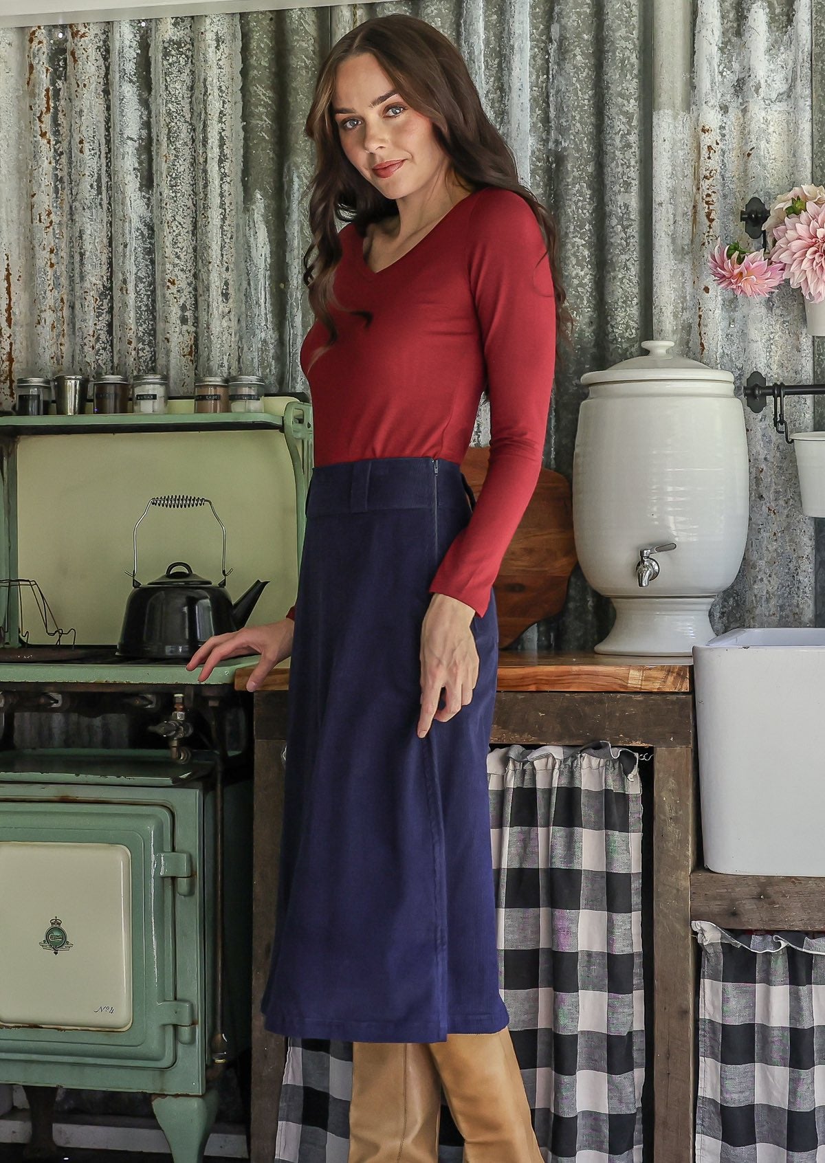 Woman standing in a rustic kitchen in a blue cotton corduroy skirt paired with a red long sleeve top and tan boots
