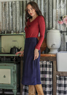 Woman standing in a rustic kitchen in a blue cotton corduroy skirt paired with a red long sleeve top and tan boots