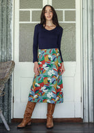 Woman wearing a colorful floral skirt and brown boots standing in front of a white door.