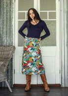 Woman poses in a colorful floral skirt and brown boots standing in front of a white door