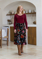 Woman wearing a cotton velvet skirt in black based multi-coloured floral print standing in a kitchen. 