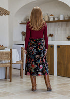 Woman standing backwards in a black based multi-coloured florals cotton velvet skirt paired with a maroon long sleeve top and tan boots