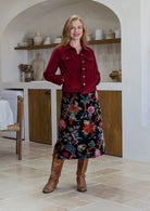 Woman in a corduroy jacket and floral cotton velvet skirt standing in a kitchen.