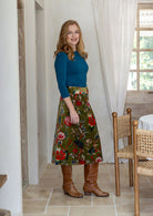 Woman standing sideways in a rustic kitchen in a moss green based multi-coloured florals cotton velvet skirt paired with a basic long sleeve top and tan boots