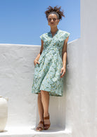 Woman stands in sage green based floral print cotton dress with box pleats, v neckline and cap sleeves 