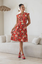Woman stands in red based floral print cotton dress with box pleats, v neckline and cap sleeves 