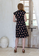 Woman stands backwards wearing 100% cotton retro style knee length dress with box pleats, cap sleeves and V neckline.