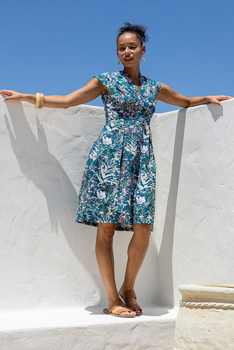 Woman stands, wearing a teal based floral cotton dress with side zip standing against a white wall and a blue sky.