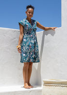 Woman stands in teal based floral print cotton dress with box pleats, v neckline and cap sleeves 