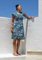 Woman standing in a living room wears knee length 100% cotton dress with delicate multicoloured wildflowers on a teal base.