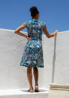 Woman stands backwards wearing teal based floral cotton dress with cap sleeves, box pleats and V neckline