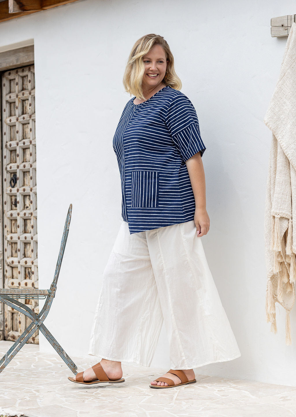 Woman stands sideways wears a navy and white striped 100% cotton relaxed fit top with an asymmetrical hem line and front pockets. 