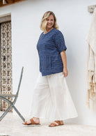 Woman stands sideways wears a navy and white striped 100% cotton relaxed fit top with an asymmetrical hem line and front pockets. 