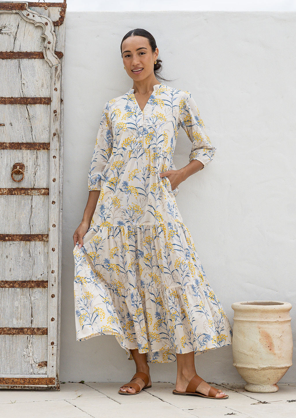 Woman wearing a 100% cotton neutral based floral maxi dress against a white wall with a rustic wooden door.