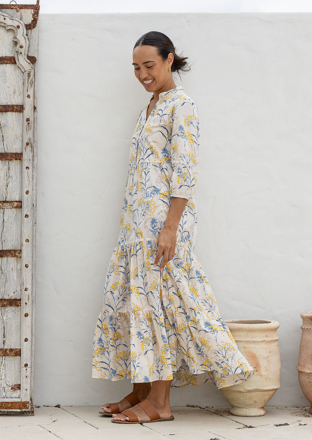 Woman stands side ways wearing a 100% cotton tiered maxi dress in neutral based floral print with 3/4 sleeves, pockets and mandarin collar