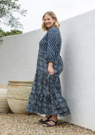 Woman stands sideways wearing a 100% cotton tiered maxi dress in floral print with 3/4 sleeves, pockets and mandarin collar