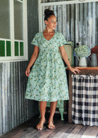 Woman wearing a pale green cotton floral button through dress with short sleeves and a V-neckline, standing in a rustic room