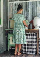 Woman in a pale green cotton floral dress standing in a rustic kitchen showing the back of the dress cinched with shoestring ties