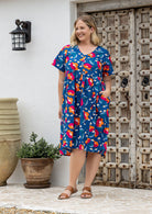 A woman modeling a short-sleeve V-neck cotton midi dress with a floral and floral print on a royal blue background, standing against a wooden door.