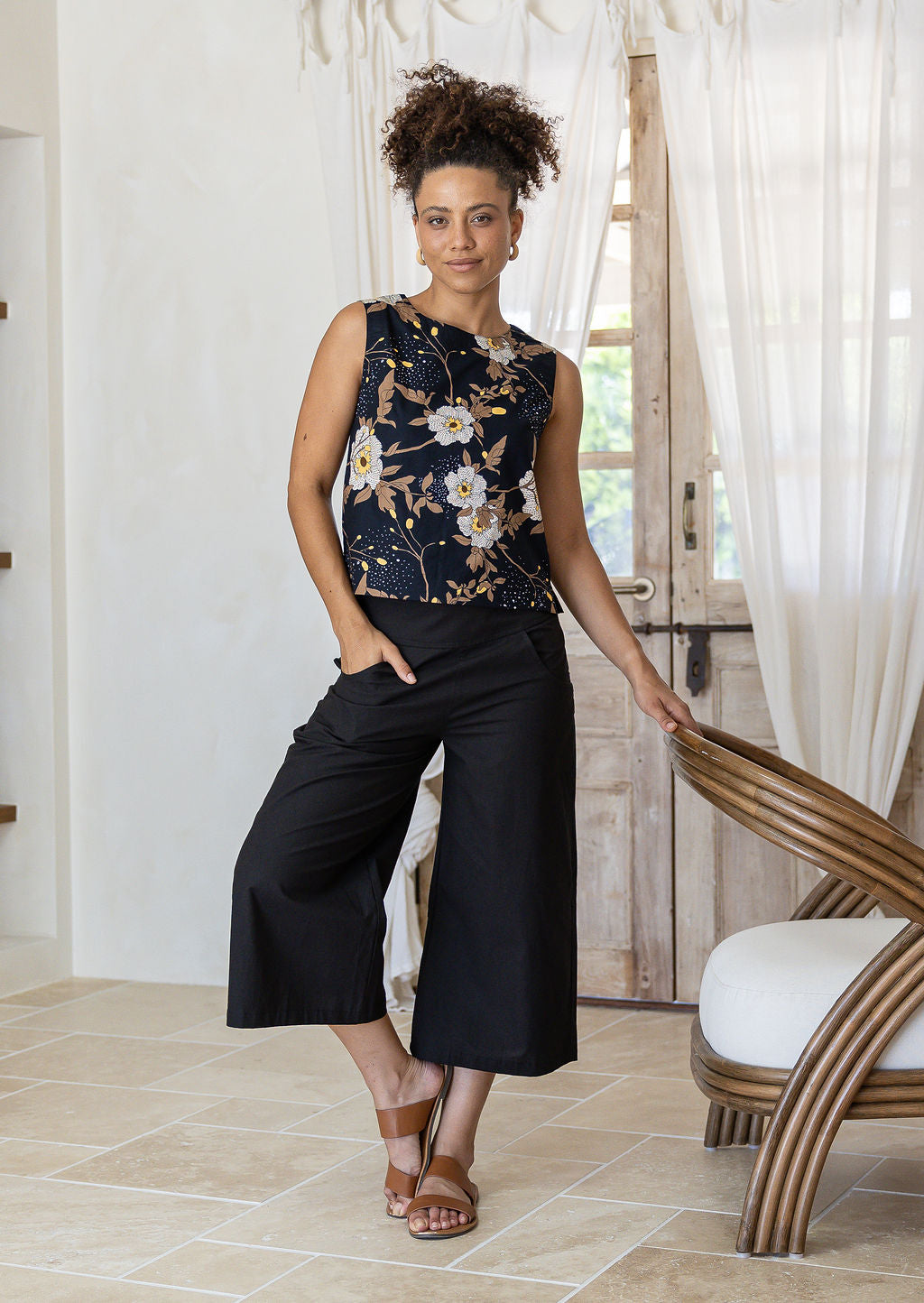 Model poses in sleeveless cotton top in black based floral print with high round neckline and side bust darts.