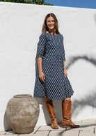 Woman in a navy blue patterned dress and brown boots standing next to a white wall