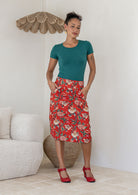 Woman wearing a knee length floral print skirt with piped detailing at the waistband and pockets, paired with a jade green rayon top